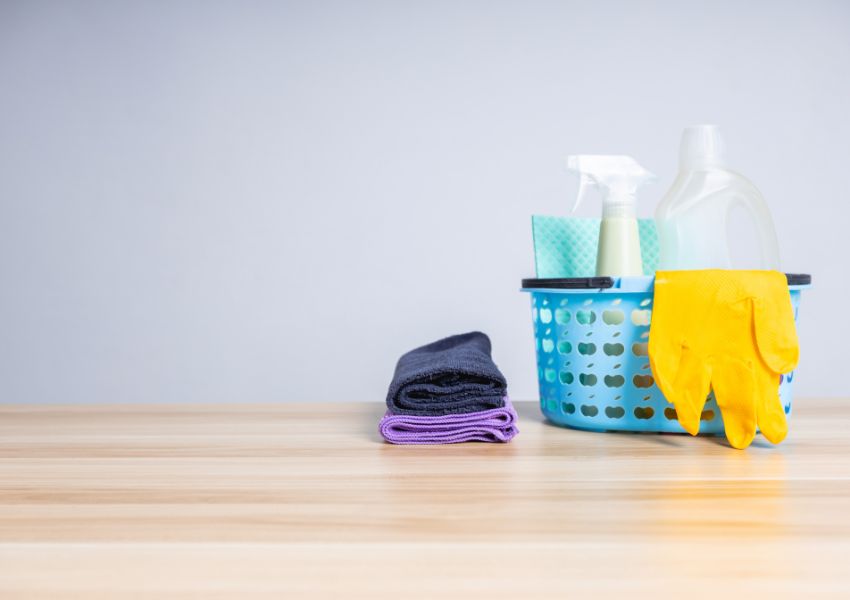 Cleaning supplies sit in a blue basket on a wooden table, including a spray bottle, yellow latex gloves, and purple and navy rags.