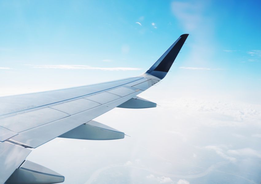 The window view of a plane is pictured with the plane's silver wing and a blue and cloudy sky.